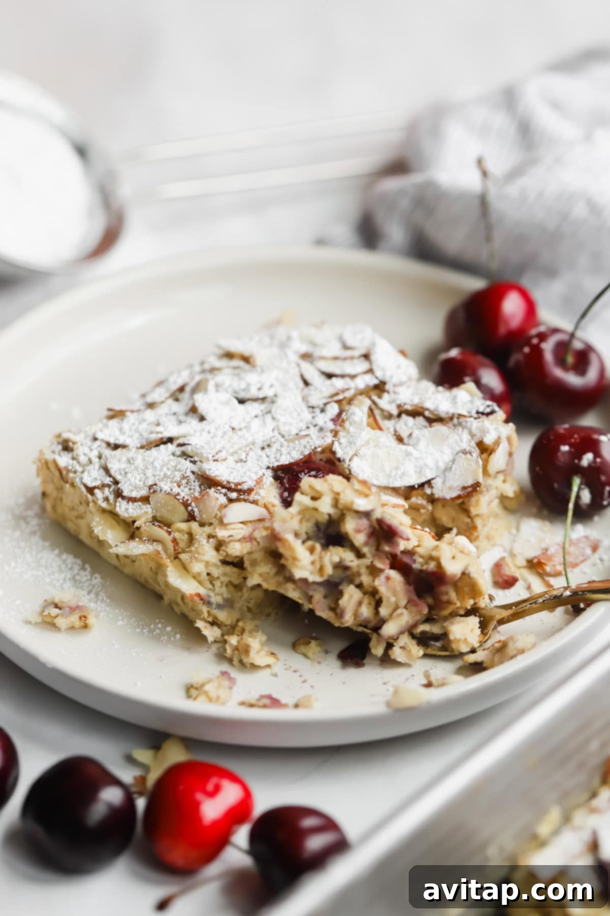 A partially eaten slice of almond and cherry cake, generously topped with powdered sugar, rests on a plate. Fresh, ripe cherries are elegantly arranged on the side, highlighting the natural ingredients of the dish.