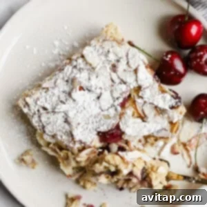 A square slice of almond cherry cake topped with powdered sugar on a plate, served with fresh cherries on the side.