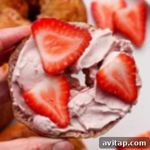 Creamy Strawberry Cottage Bagels 12 A hand holds a bagel half topped with pink cream cheese and sliced strawberries, with more strawberries and bagels on a plate in the background.
