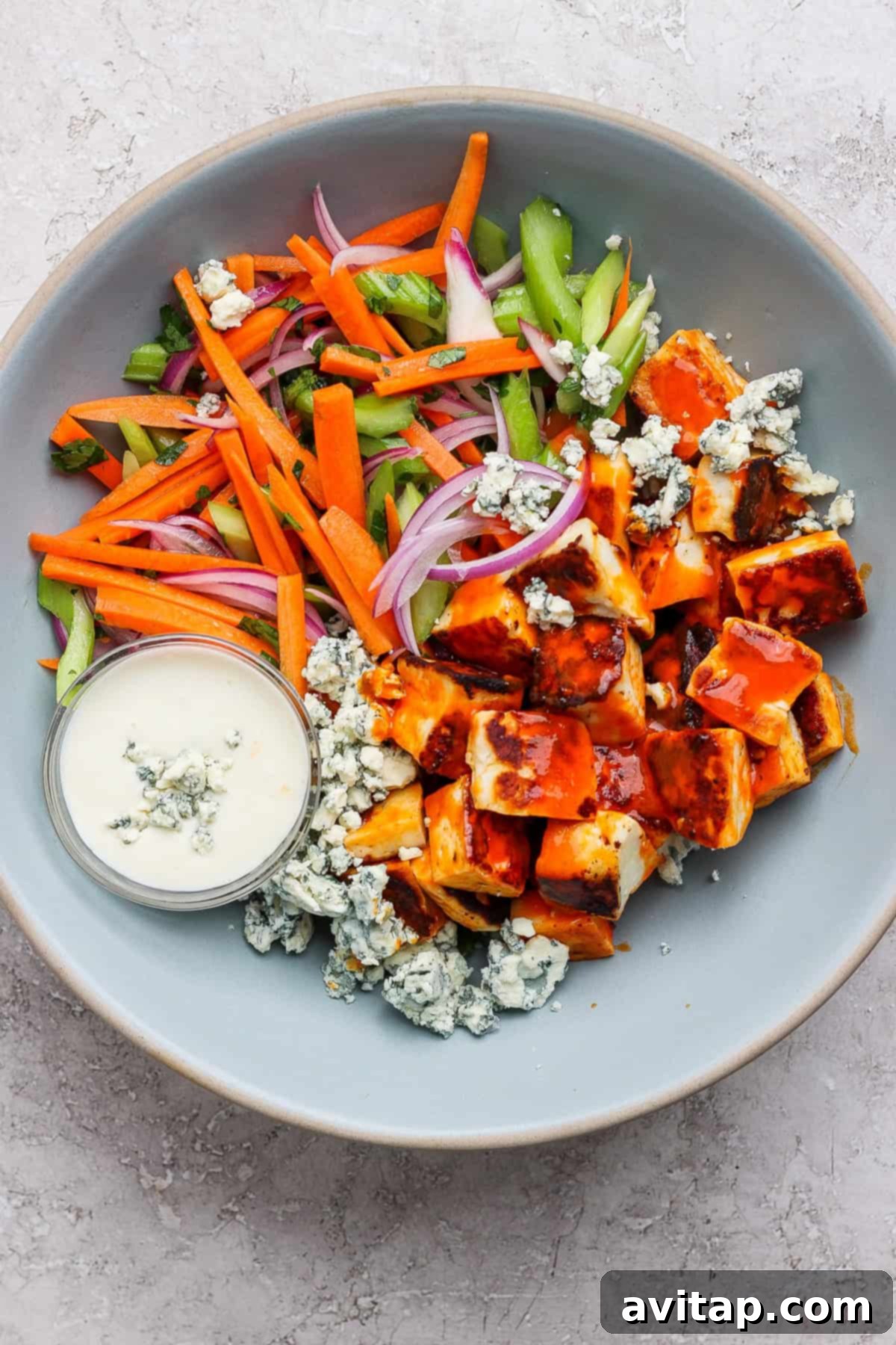 A salad bowl with cubed grilled chicken, crumbled blue cheese, carrot sticks, celery, red onion, and a small dish of creamy dressing on the side.