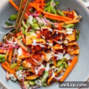 A salad with grilled chicken pieces, carrots, celery, red onion, blue cheese crumbles, drizzled with dressing, in a large bowl with two spoons.