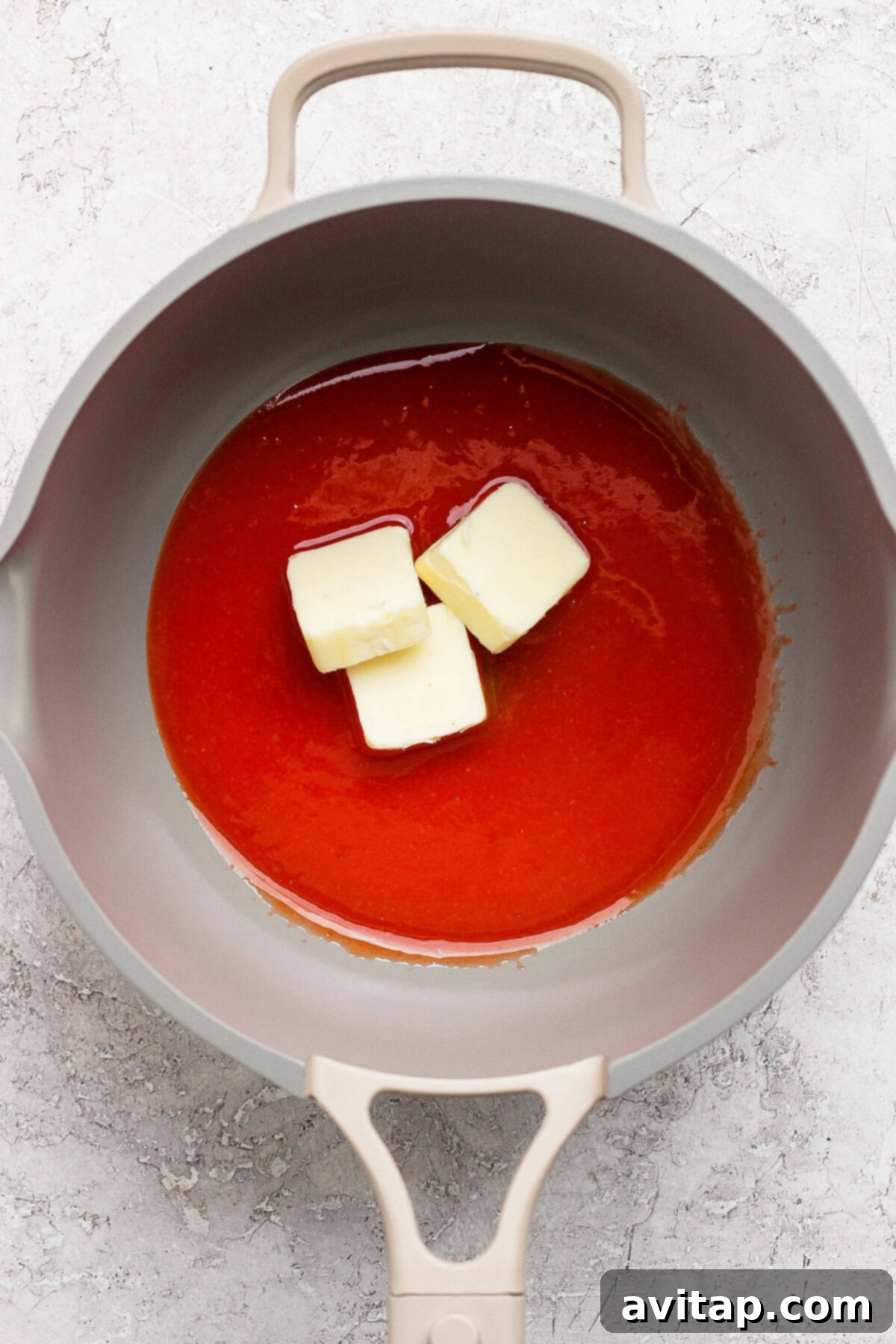 A grey pan contains a red liquid with three chunks of butter melting in it. The background is a light, textured surface.