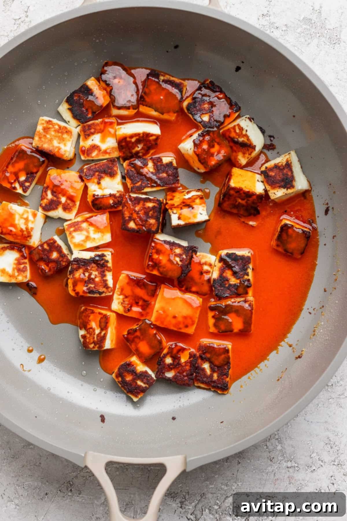 Pan-fried paneer cubes coated in a rich red sauce in a non-stick pan on a light gray surface.