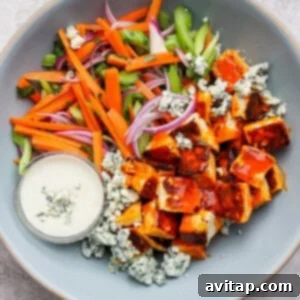 A bowl with diced carrots, celery, red onion, blue cheese crumbles, and buffalo sauce-coated tofu. A small cup of creamy dressing is on the side.