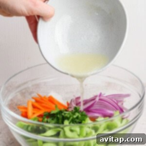 Person pouring a light dressing into a bowl of chopped vegetables including carrots, red onions, celery, and herbs.