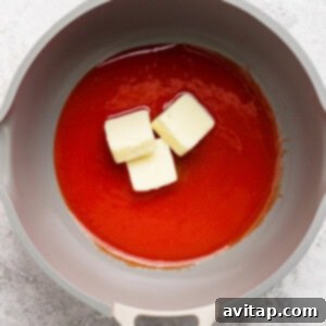 A grey pan contains a red liquid with three chunks of butter melting in it. The background is a light, textured surface.