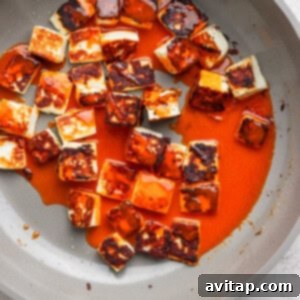 Pan-fried paneer cubes coated in a rich red sauce in a non-stick pan on a light gray surface.