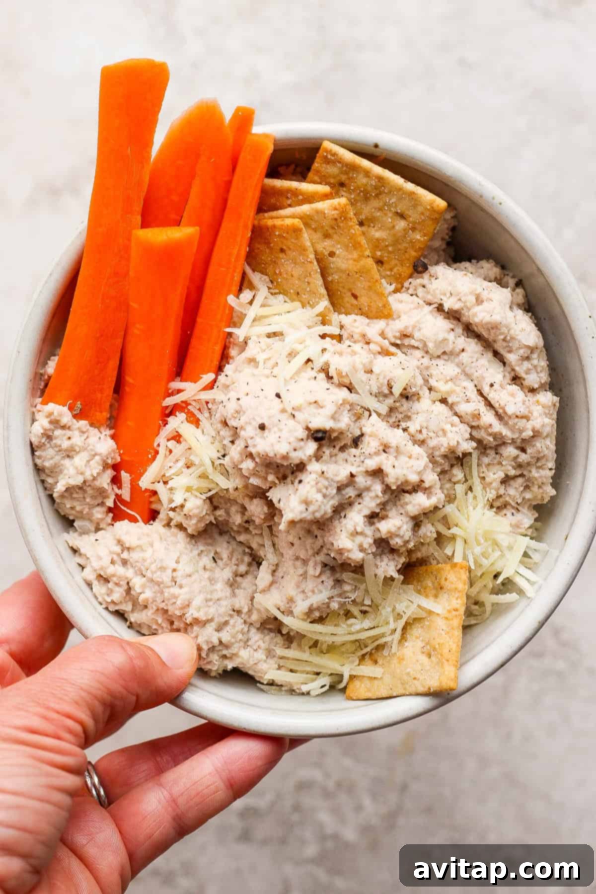 A hand holds a rustic wooden bowl containing crisp carrot sticks, an assortment of savory crackers, a sprinkle of shredded cheese, and a generous scoop of creamy, flavorful chicken Caesar salad spread, highlighting its perfect texture and appetizing presentation.
