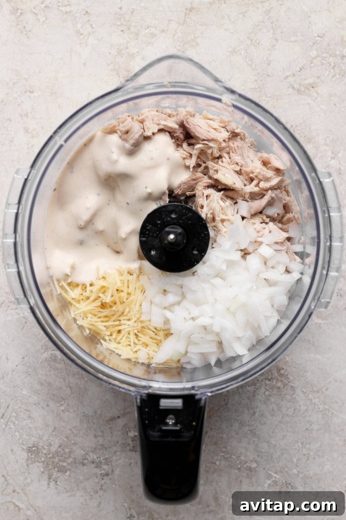 From a bird's-eye view, a food processor bowl showcases a tempting medley of shredded chicken, finely chopped red onions, a generous scattering of grated Parmesan cheese, and a rich, creamy sauce, all prepped and ready to be blended into a savory chicken Caesar salad spread.