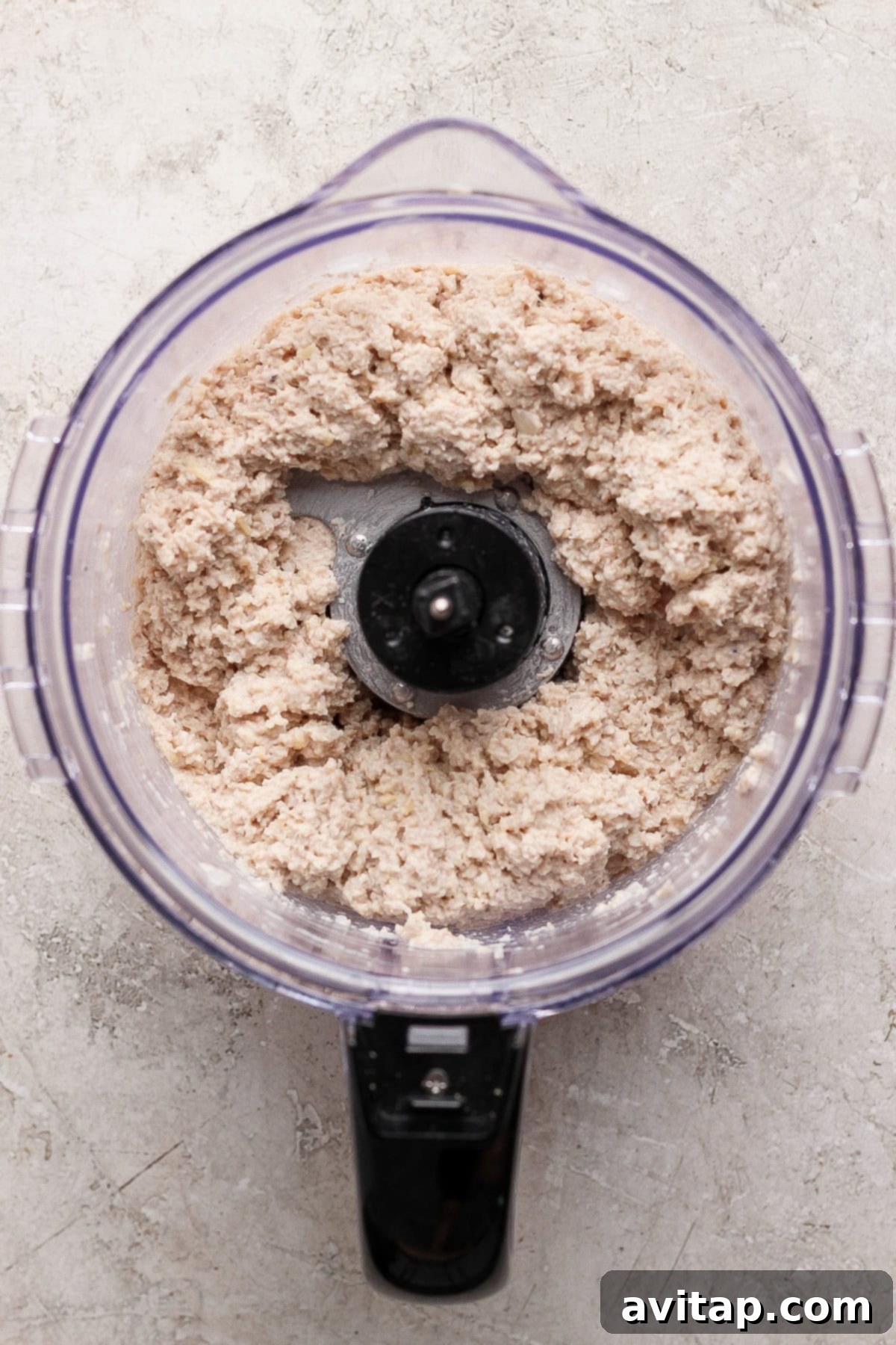 A high-angle view captures a food processor filled with a freshly prepared, coarse, pale chicken Caesar salad spread, resting on a subtly textured, light-colored kitchen counter, showcasing its perfectly blended consistency.