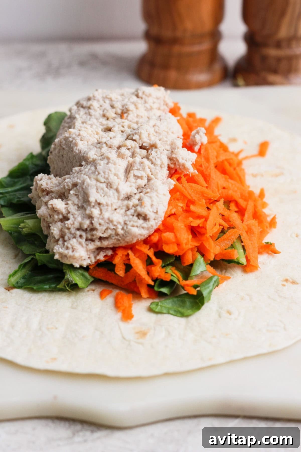 A soft flour tortilla is neatly arranged on a pristine white surface, laden with a vibrant mix of crisp leafy greens, finely grated carrots, and a generous, creamy scoop of either tuna or chicken Caesar salad spread, inviting a healthy and flavorful bite.