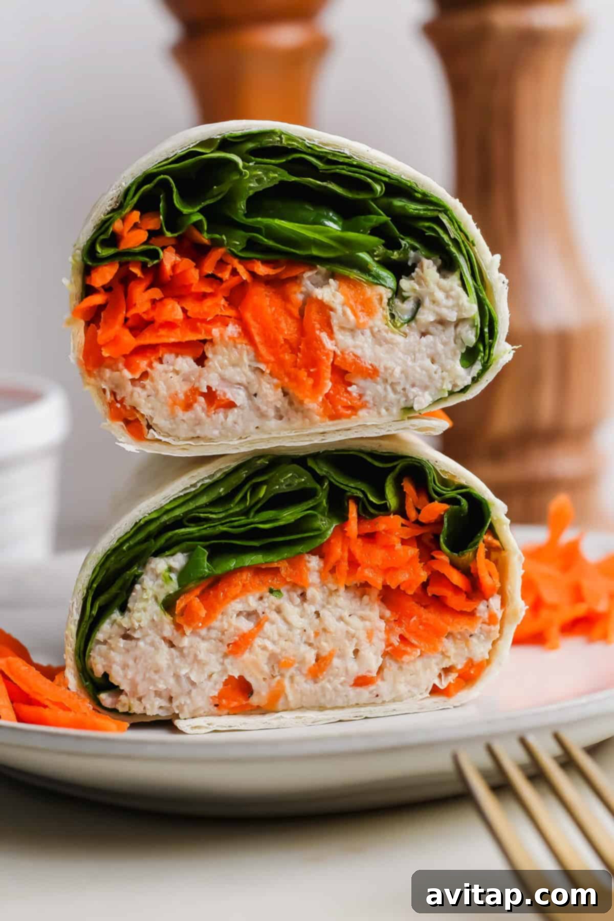 A vibrant, halved wrap revealing layers of finely shredded carrots, fresh leafy greens, and a creamy chicken salad mixture, elegantly stacked on a pristine white plate, with a silver fork resting invitingly beside it.