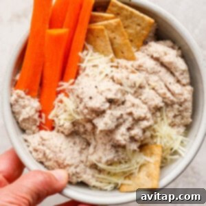 A bowl containing crisp carrot sticks, crackers, shredded cheese, and a creamy, inviting chicken Caesar salad dip, held gently in one hand.