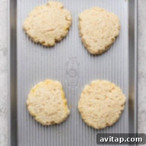 Four unbaked biscuit dough rounds are spaced evenly on a metal baking sheet placed on a light-colored surface.