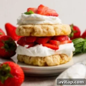 A strawberry shortcake dessert with layers of biscuits, whipped cream, and sliced strawberries on a plate, surrounded by whole strawberries and mint leaves.