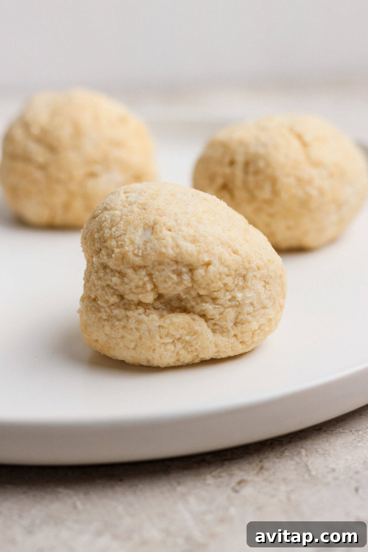 Three round, lightly textured dough balls are arranged on a white plate with a neutral background.