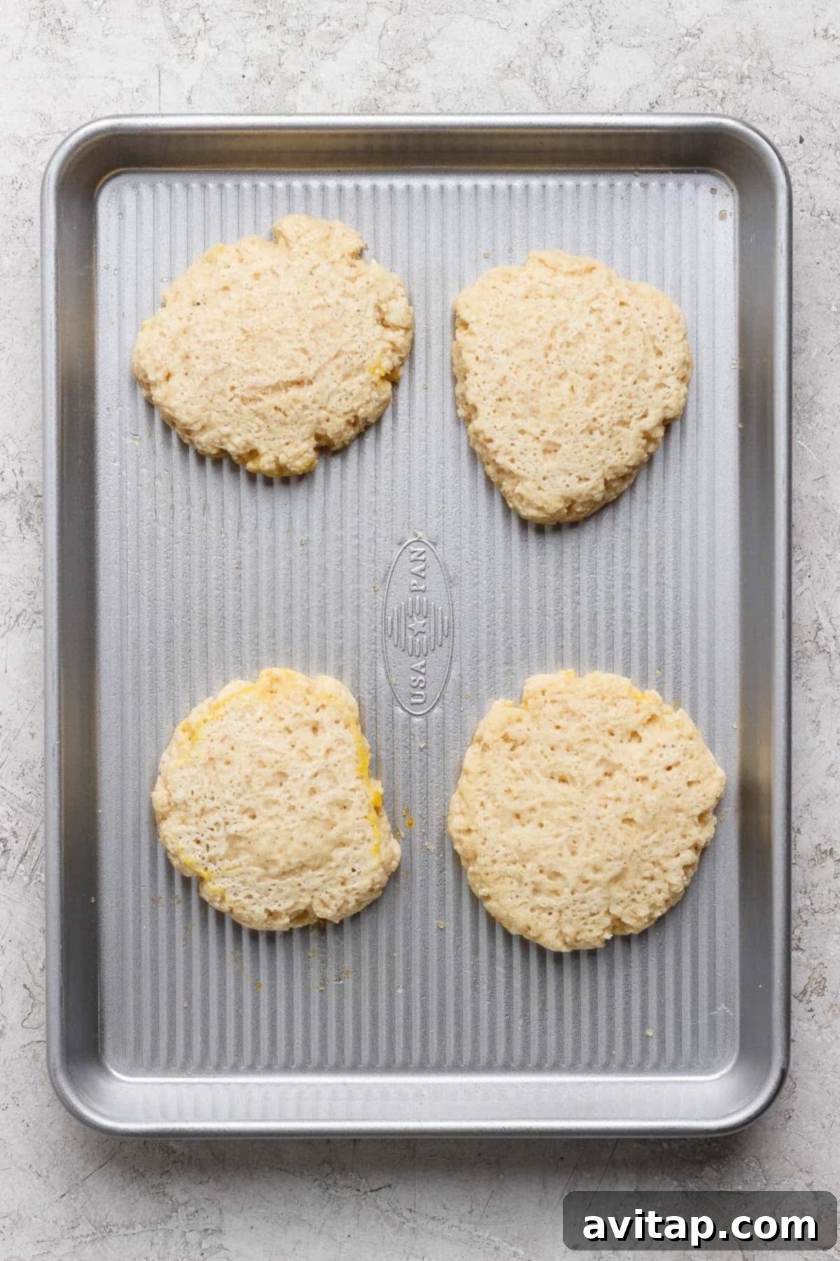Four unbaked biscuit dough rounds are spaced evenly on a metal baking sheet placed on a light-colored surface.