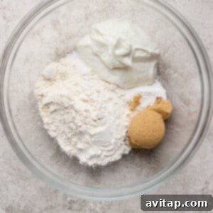 A glass bowl containing flour, brown sugar, and a scoop of white yogurt on a light textured surface.