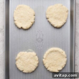 Four unbaked biscuit dough rounds evenly spaced on a silver baking sheet, placed on a light-colored surface.