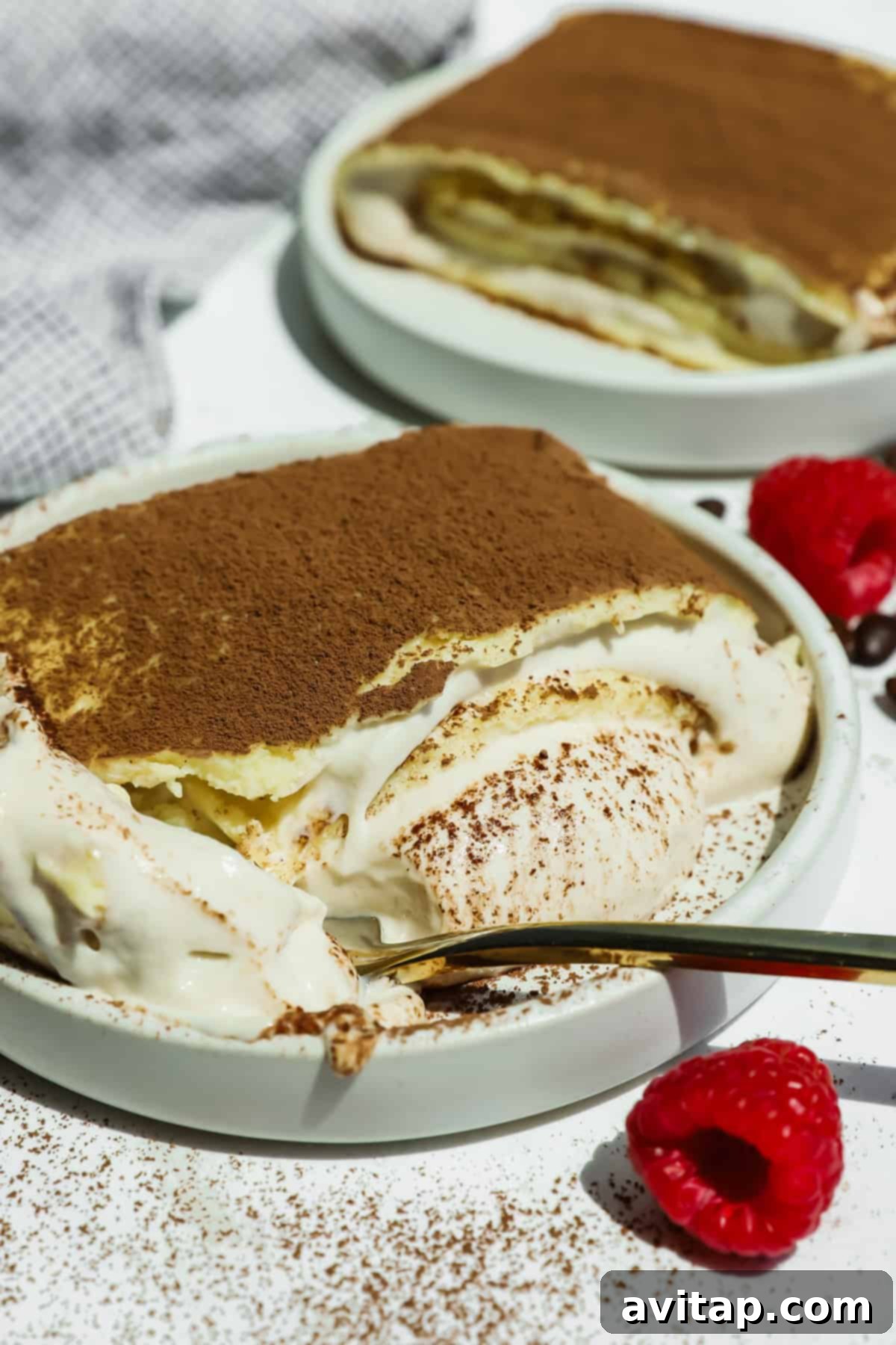 A plate of cottage cheese tiramisu crepe roll with a portion scooped out reveals creamy layers, dusted with cocoa powder. Fresh raspberries and a gold spoon are nearby.