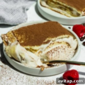 A plate of cottage cheese tiramisu crepe roll with a spoonful taken out, topped with cocoa powder, sits next to fresh raspberries on a white surface.