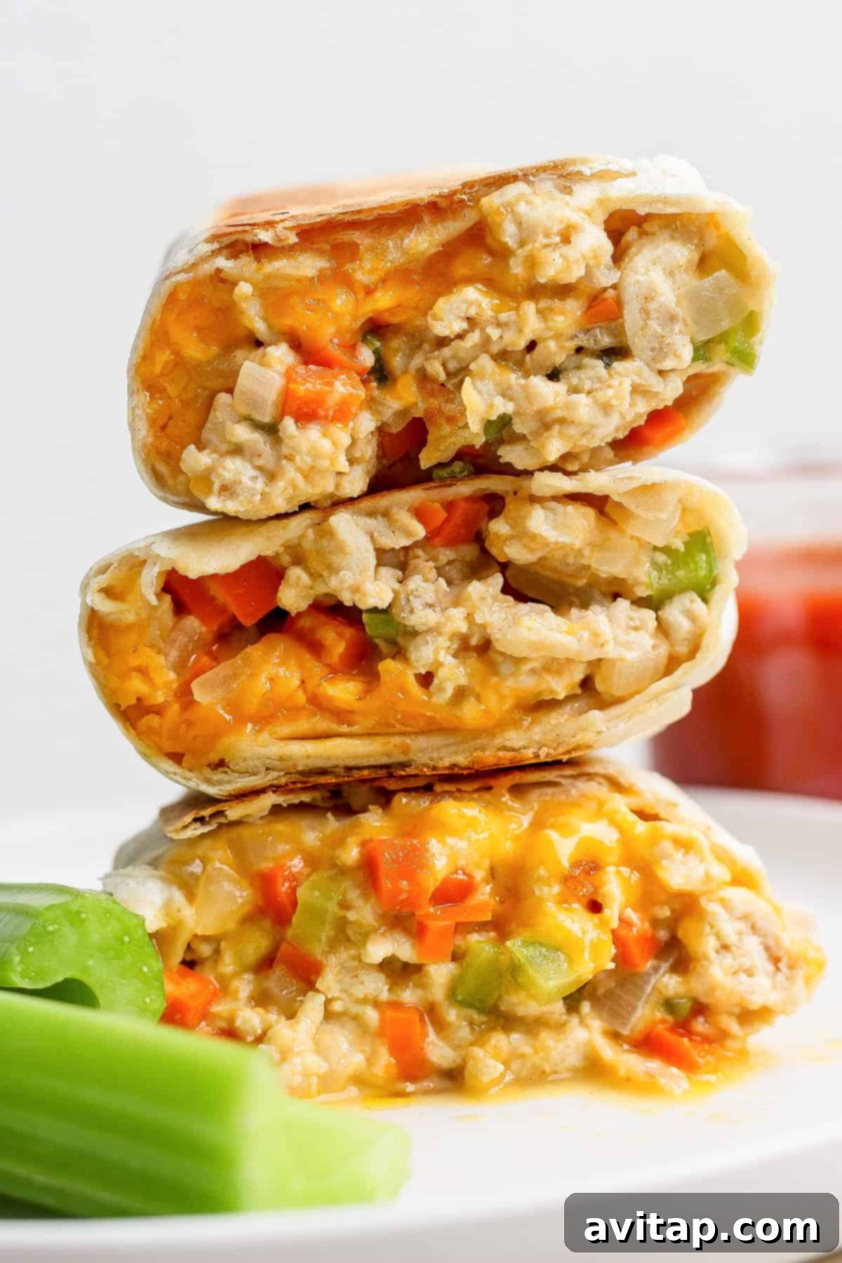 Three halves of a burrito filled with scrambled eggs, cheese, and diced vegetables are stacked on a plate next to celery sticks.