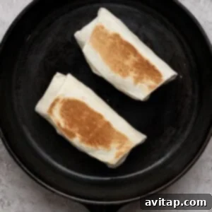 Two folded burritos being toasted in a black cast iron skillet on a light textured surface.