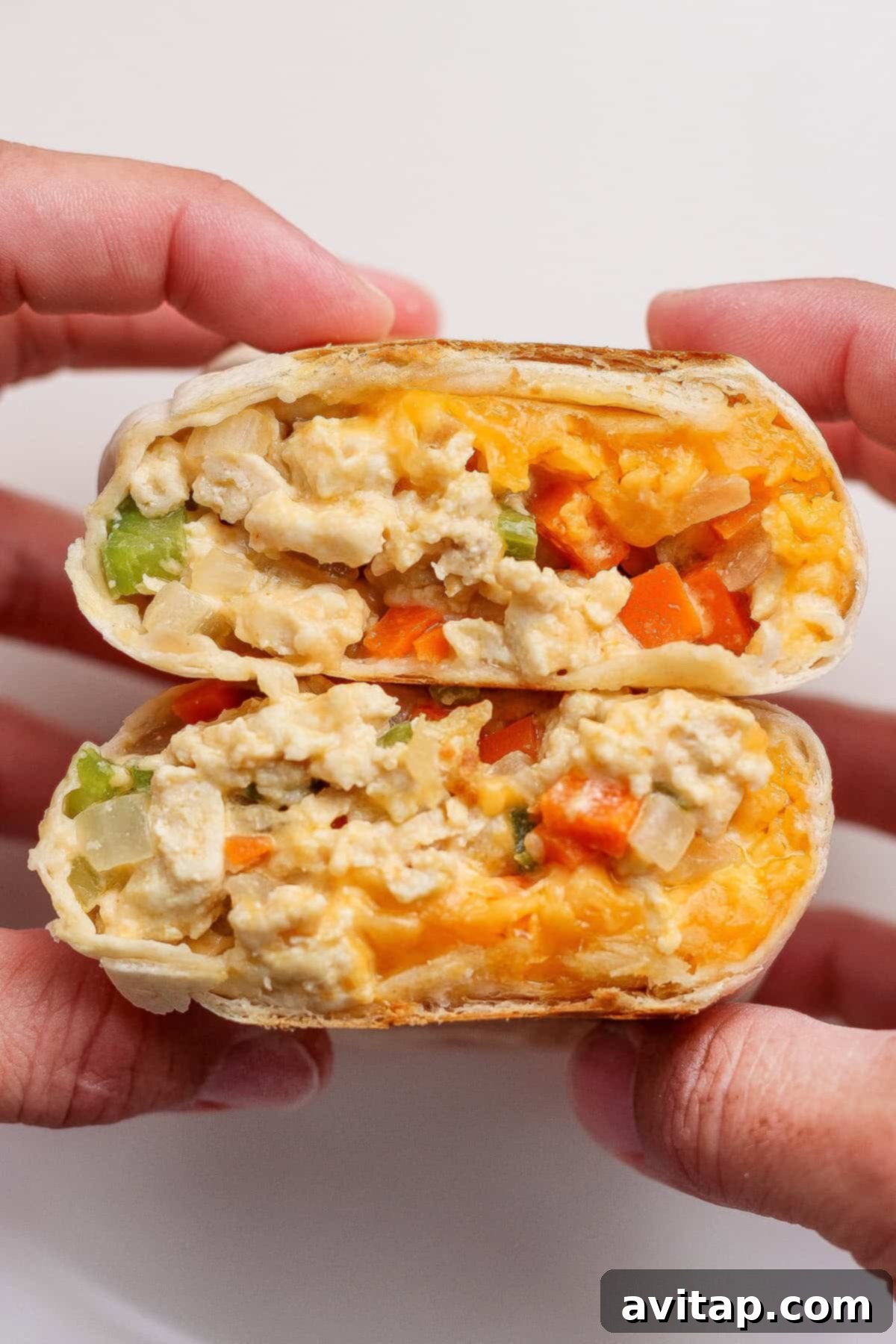 Two hands hold a breakfast burrito cut in half, showing scrambled eggs, cheddar cheese, and diced vegetables inside a tortilla wrap.