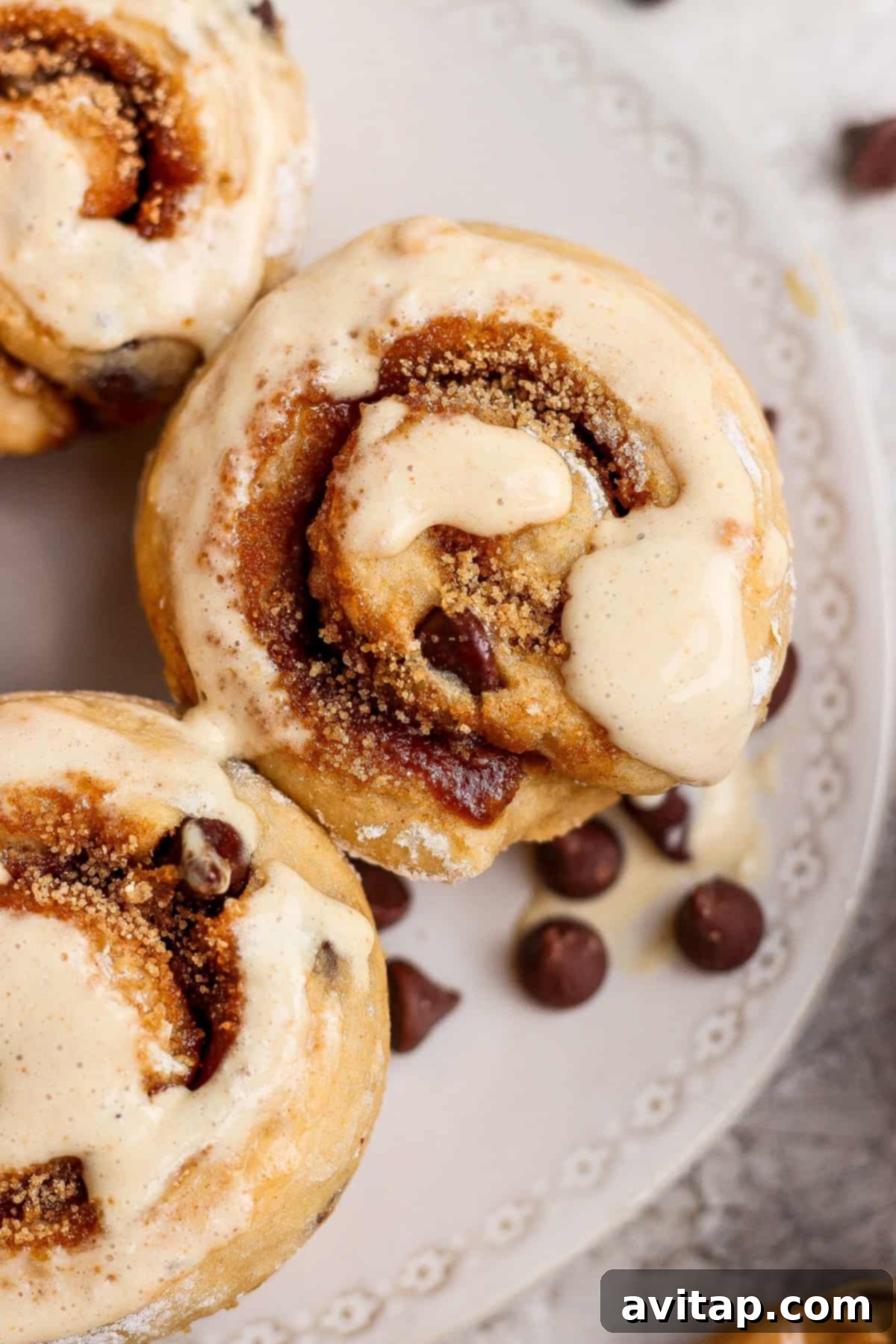 Cozy Fall Protein: Cottage Cheese Delights 4 Close-up of cinnamon rolls topped with icing on a decorative white plate, with several chocolate chips scattered nearby, showcasing a high-protein dessert.