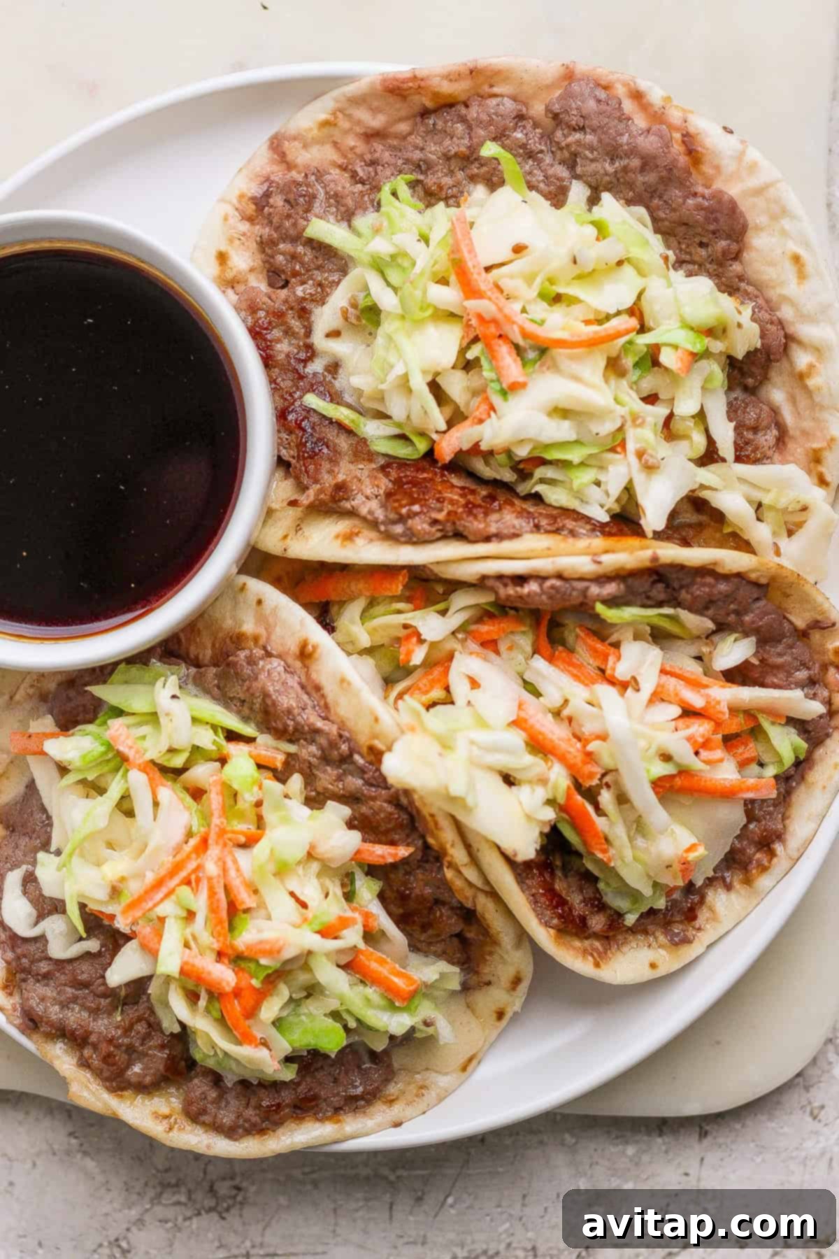 Three smash burger tacos filled with seasoned beef, shredded cabbage, and carrots, served on a white plate with a small bowl of dark dipping sauce.