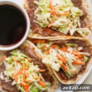 Three tacos filled with refried beans, shredded cabbage, and carrots are served on a white plate with a small bowl of dark dipping sauce.