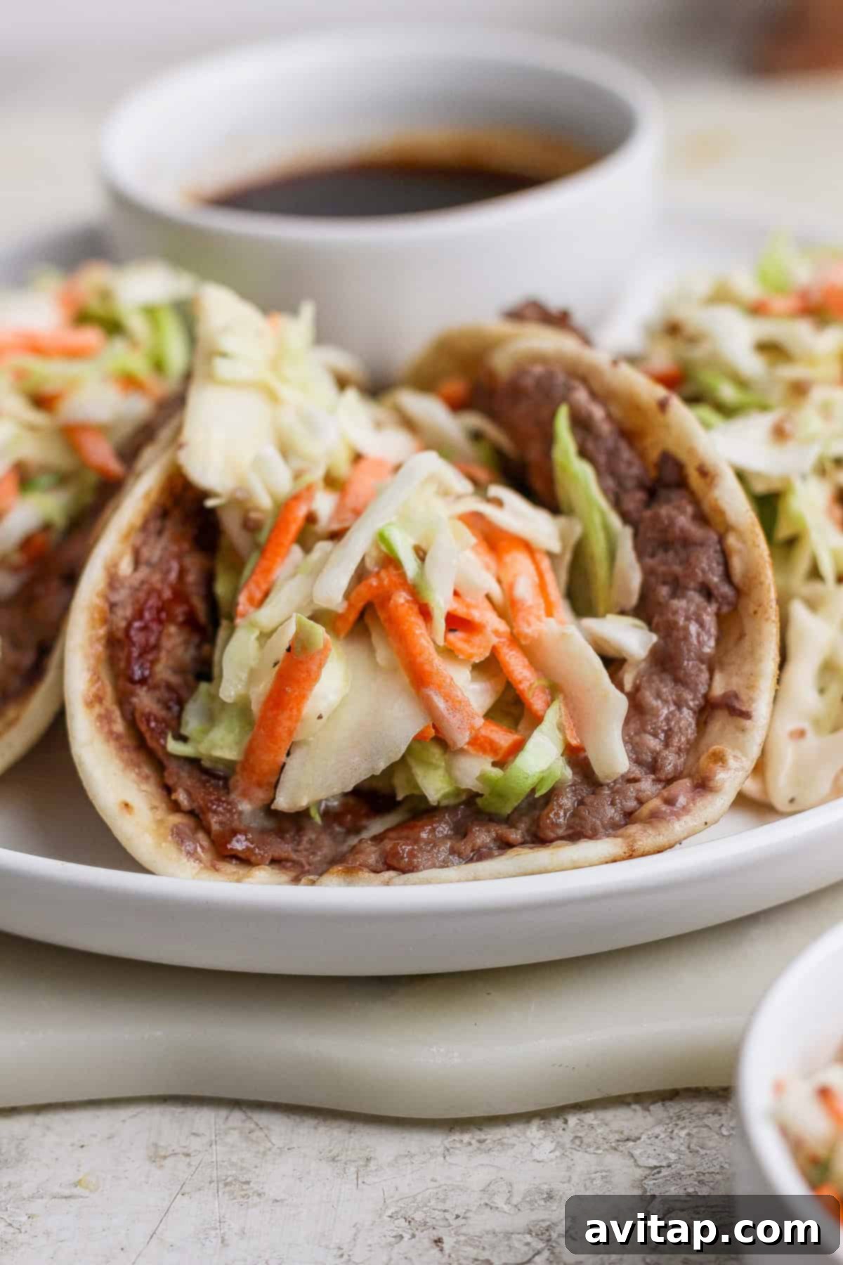 A soft taco filled with cooked beef, shredded cabbage, and carrots, served on a white plate with a small bowl of sauce in the background.