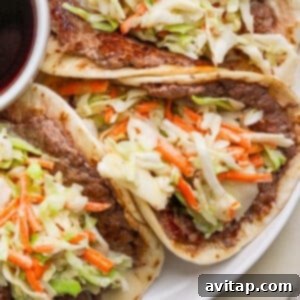 Three tacos filled with cooked beef and topped with shredded cabbage and carrots, served on a plate next to a small bowl of dark sauce.