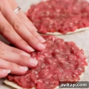 Two hands press raw ground meat onto a flatbread or dough circle, forming a thin layer. Another prepared flatbread with meat is visible in the background.