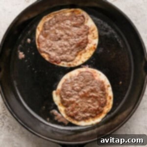 Two corn tortillas topped with ground meat cooking in a black cast iron skillet on a light-colored textured surface.