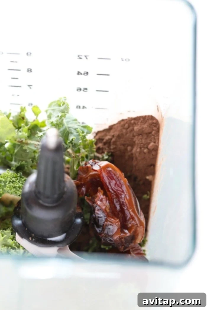 Pitted Medjool dates, fresh kale leaves, and dark chocolate cocoa powder being gathered and ready to be added to a high-speed blender for a nutritious protein smoothie.