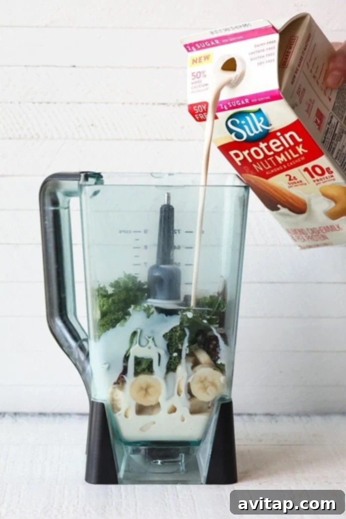 Pouring Silk Protein Nut Milk into a high-speed blender already containing frozen bananas, dates, kale, and cocoa powder for a protein smoothie.