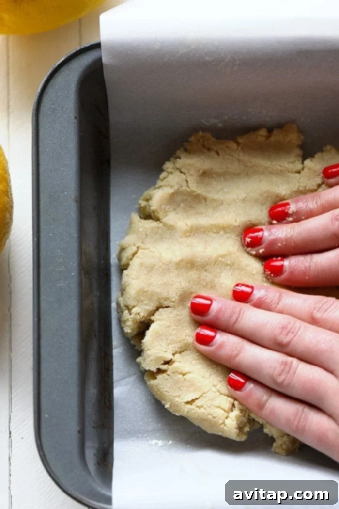 Hands pressing a homemade lemon bar crust into a baking pan, showing the even distribution.