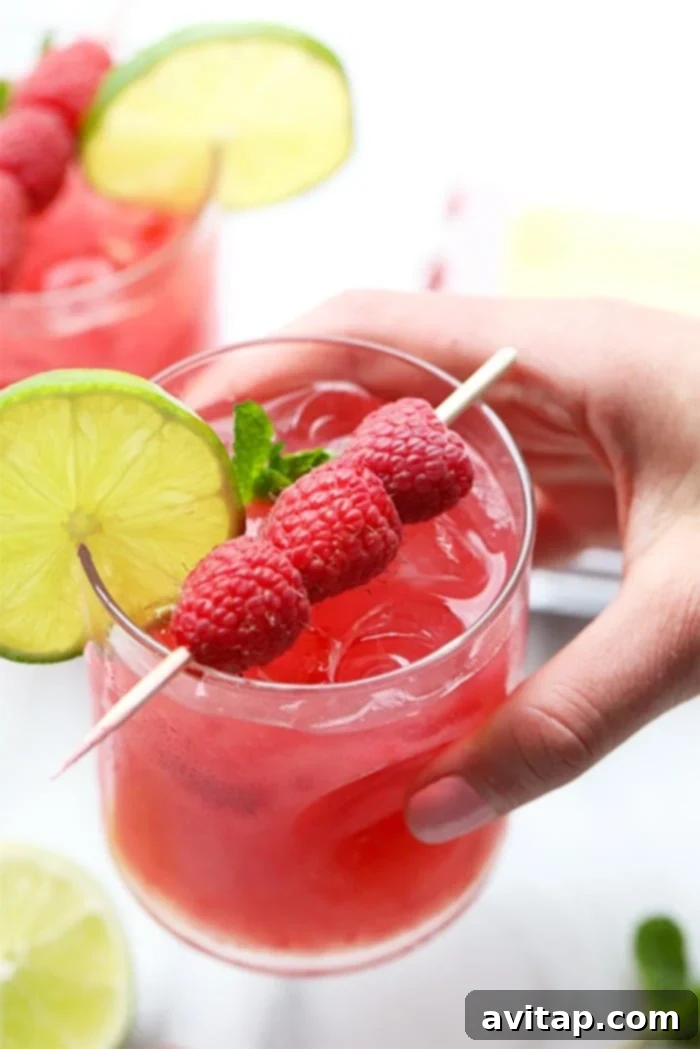 A hand holding a vibrant Ginger Raspberry Mojito garnished with fresh raspberries and a lime wedge.