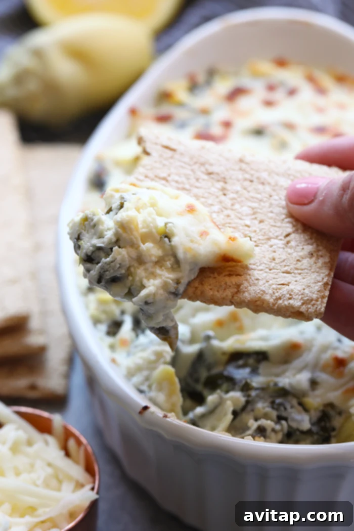Enjoy this AMAZING lightened-up, restaurant-style Healthy Spinach and Artichoke Dip for a nutrition-packed snack or appetizer all year round. This spinach and artichoke dip will quickly become a crowd-pleasing recipe you'll make again and again. A cracker being dipped into a bowl of lightened-up spinach artichoke dip, highlighting its creamy texture.
