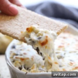 A person is dipping a cracker into a bowl of Lightened Up Spinach and Artichoke Dip.