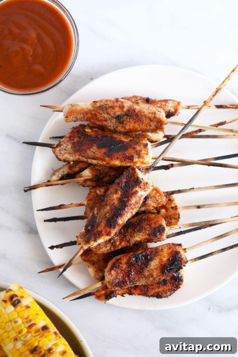 Grilled skewers on plate next to BBQ sauce and grilled corn. 