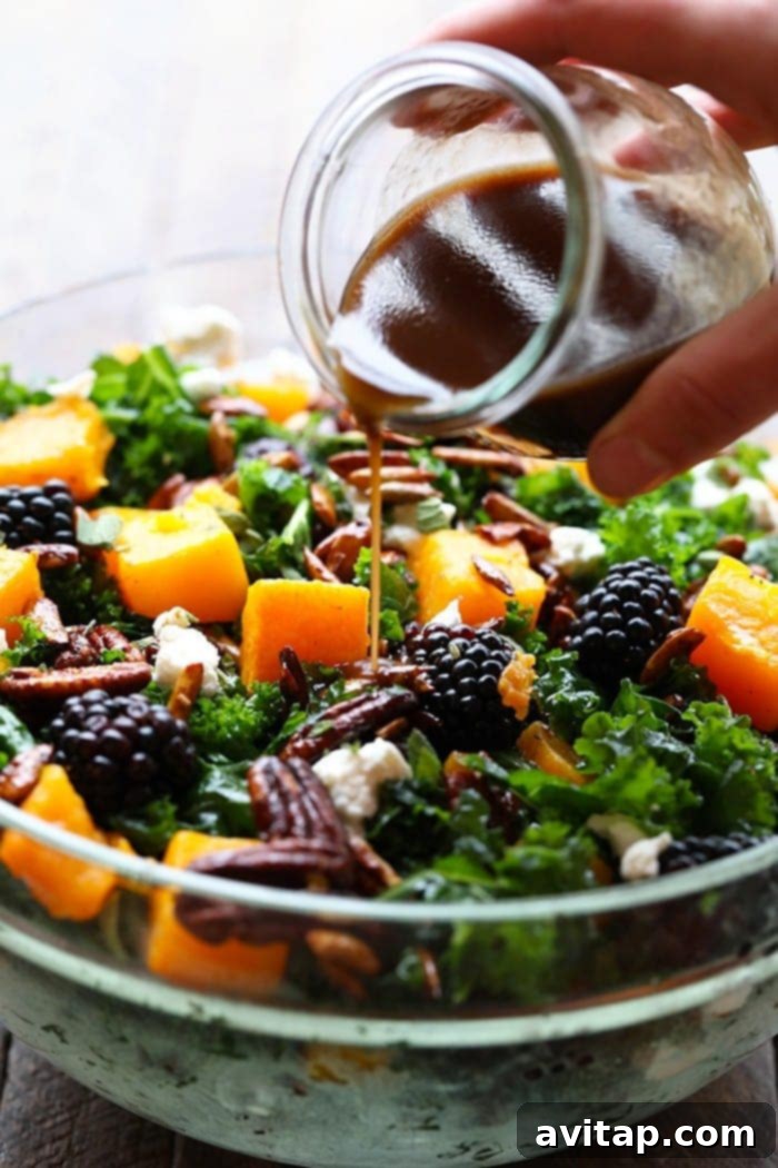 Hand pouring homemade balsamic dressing from a glass jar onto a large salad bowl filled with kale, butternut squash, and blackberries.