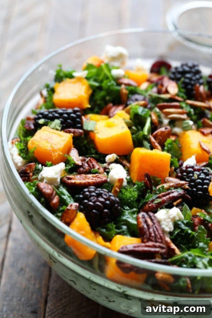 A large, rustic bowl filled with a colorful harvest salad, featuring massaged kale, roasted butternut squash, blackberries, candied pecans, goat cheese, and dried cranberries, ready to serve.