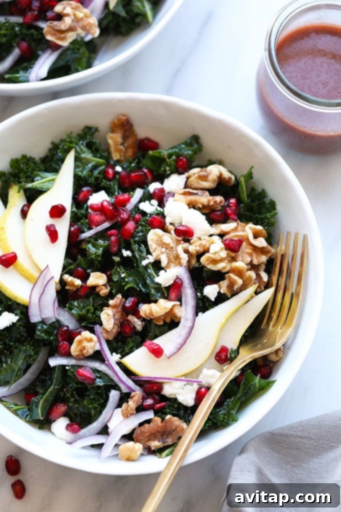 A beautifully presented kale salad in a large bowl, featuring pomegranate arils, pear slices, walnuts, and goat cheese, with a rich red pomegranate dressing