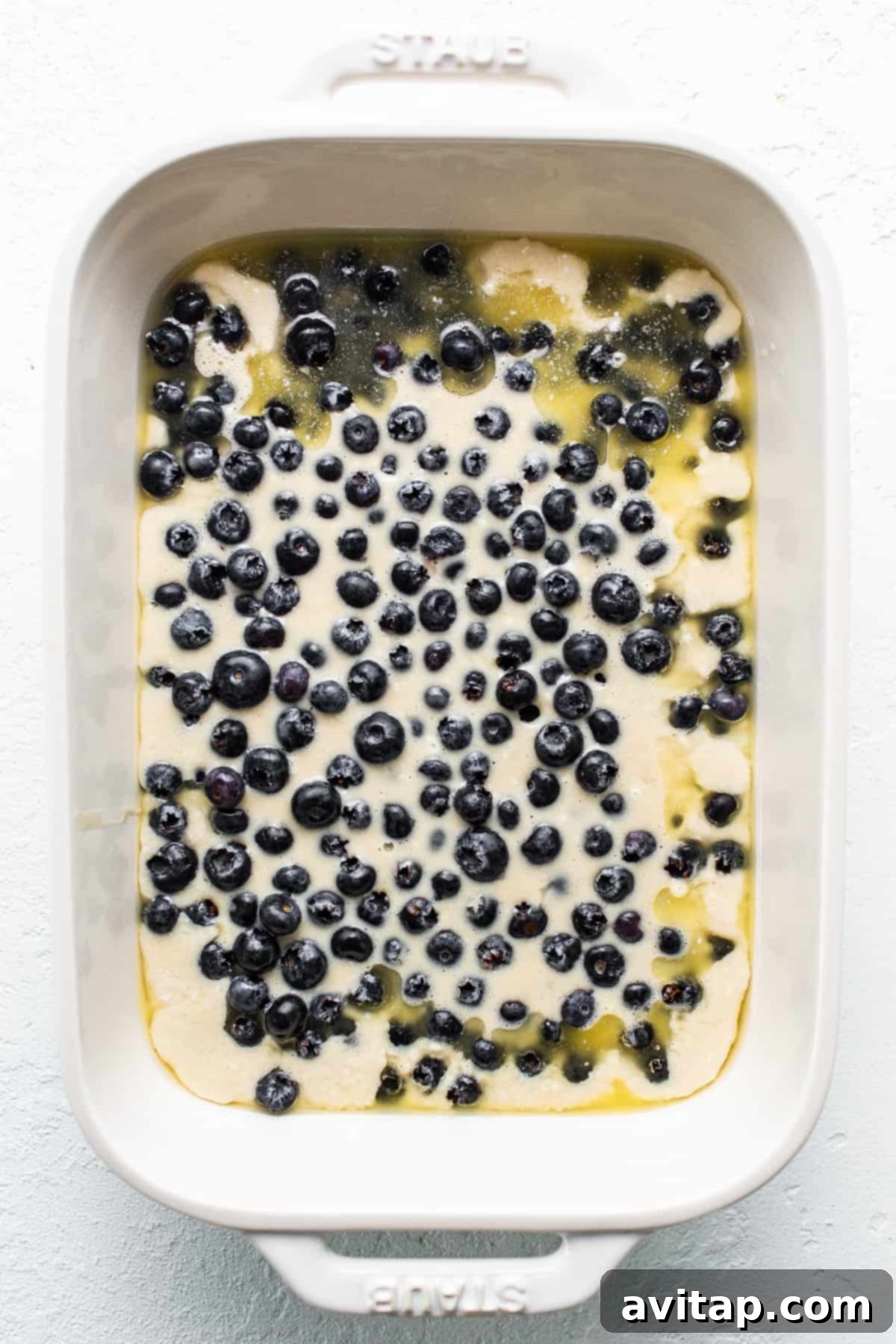 An uncooked blueberry cobbler in a clear glass baking dish, showing the vibrant blue berries scattered over the light-colored batter, ready for the oven.