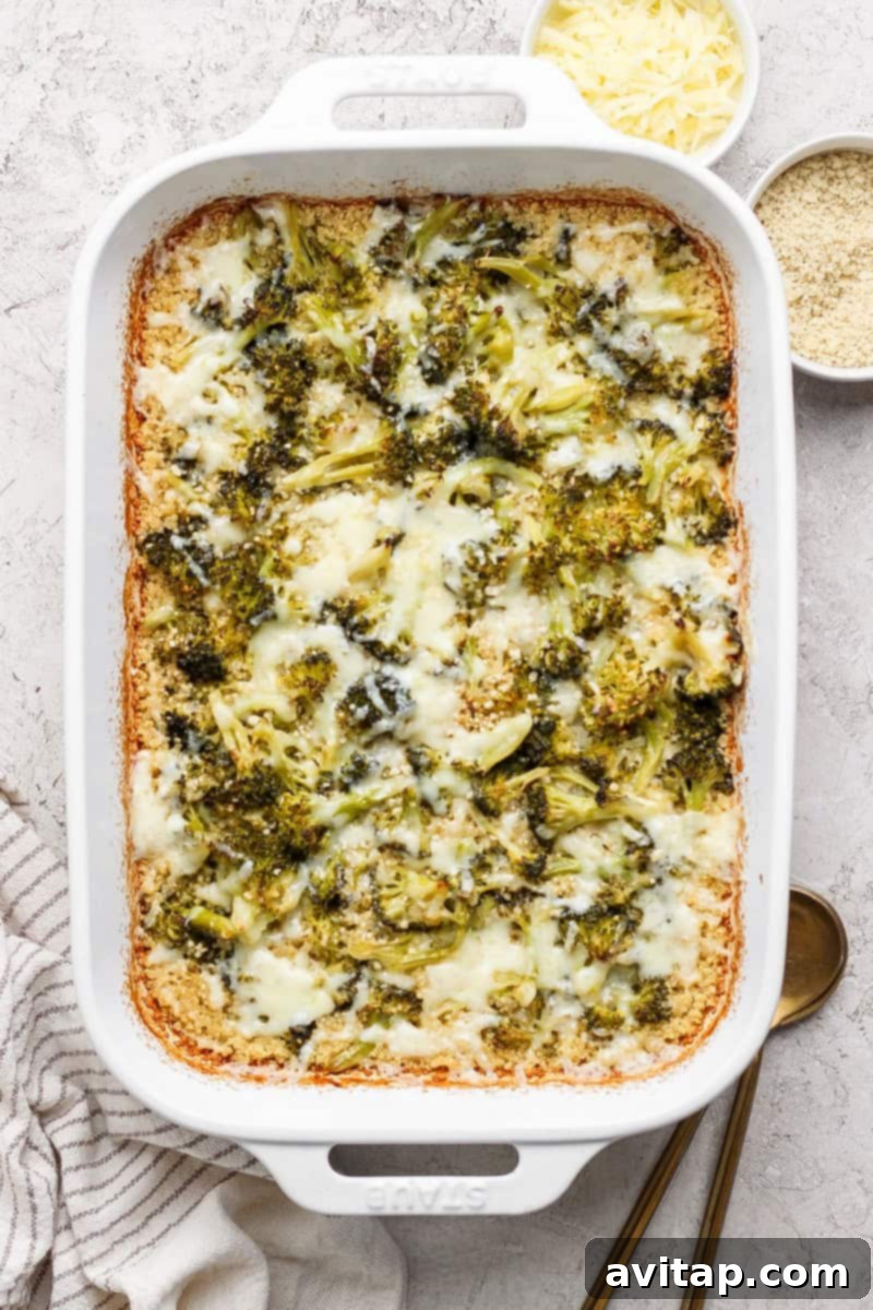 Baked broccoli casserole with melted cheese in a white dish, next to bowls of grated cheese and breadcrumbs on a textured surface.