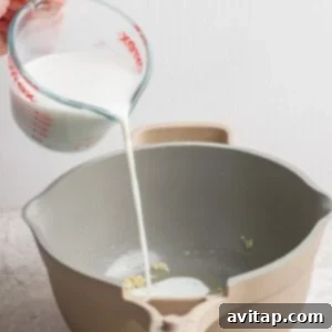 A hand pours milk from a measuring cup into a saucepan containing small food pieces, on a textured surface.