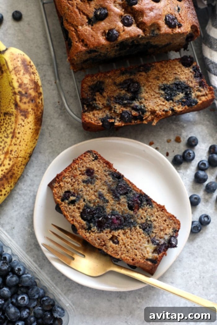 Perfectly moist blueberry banana bread made with whole grains, Greek yogurt, fresh blueberries, and maple syrup! A perfectly baked slice of blueberry banana bread on a white plate, ready to be enjoyed.