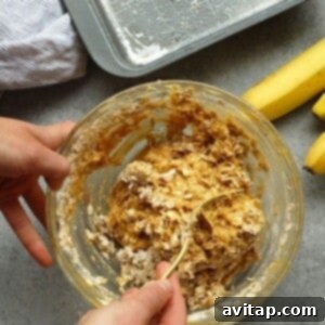 Peanut Butter Banana Oatmeal Bake 3 Person mixing banana baked oatmeal batter in a glass bowl next to a baking tray and fresh bananas on a gray counter.